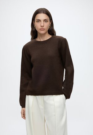 Kvinde iført en mørkebrun langærmet sweater og hvide plisserede bukser, stående mod en ensfarvet lys baggrund, kiggende fremad.