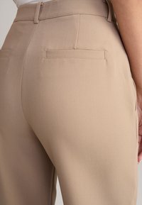 Beige maßgeschneiderte Hosen mit glatter Textur, zwei Gesäßtaschen und einer figurbetonten Passform, seitlich betrachtet.