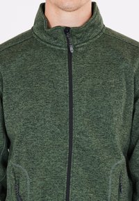 Uomo che indossa una giacca in pile verde con zip e colletto alto, cuciture visibili e tasche laterali con zip, su uno sfondo bianco semplice.