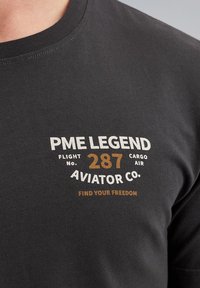 Black cotton t-shirt featuring a round neckline, with white and brown graphic text: “PME LEGEND FLIGHT NO. 287 CARGO AIR AVIATOR CO.”