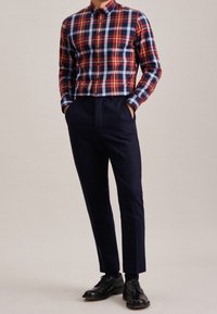Mężczyzna stojący z rękami w kieszeniach, ubrany w koszulę w czerwono-niebieską kratę, granatowe spodnie i czarne skórzane buty, na neutralnym tle.