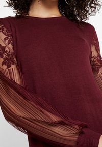 Donna che indossa un top a maglia bordeaux con maniche in pizzo trasparenti caratterizzate da motivi floreali e righe verticali.