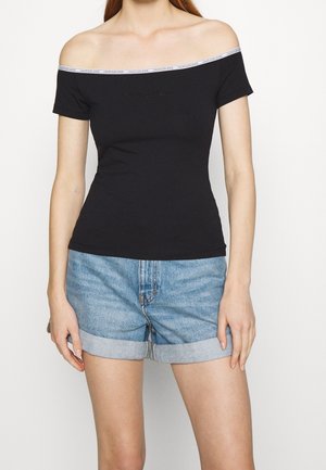 Kobieta nosząca czarny top z odkrytymi ramionami z białym wykończeniem z logo Calvin Klein, zestawiony z jasnoniebieskimi podwiniętymi szortami denimowymi.