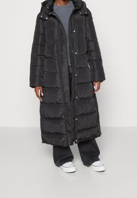 Sort pufferjakke med et langt design, der når til taljen, hætte, lynlås og trykknapper. Har quiltet tekstur og sidelommer.