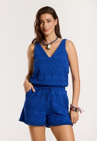 Plavi kombinezon bez rukava s teksturiranim uzorkom školjki, V-izrezom i strukom s vezicom. Završava s iznjedrenim rubovima i džepovima.