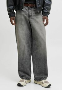 Jean en denim gris à jambe large avec une finition délavée et des coutures contrastées, associés à des baskets beiges et une veste noire.