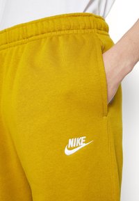 Tepláky Nike z bavlny v barvě hořčicově žluté. Mají elastický pas, boční kapsy a bílé logo na levém boku. Hladká textura.