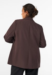 Blazer marron avec une texture lisse, fente arrière simple, col cranté et épaules tombantes, présentant une coupe décontractée et un design ajusté.