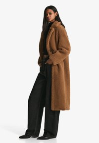 Rjav, teksturiran dolg plašč z gumbnim sprednjim delom, velikim ovratnikom in stranskimi žepi, kombiniran s črnimi širokimi kavbojkami in špičastimi čevlji.