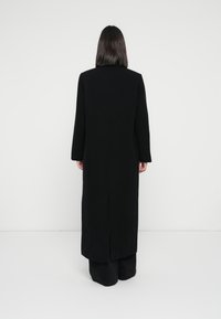 Cappotto lungo nero con taglio dritto, caratterizzato da maniche lunghe e una fessura sulla schiena. Design semplice, realizzato in un tessuto strutturato.