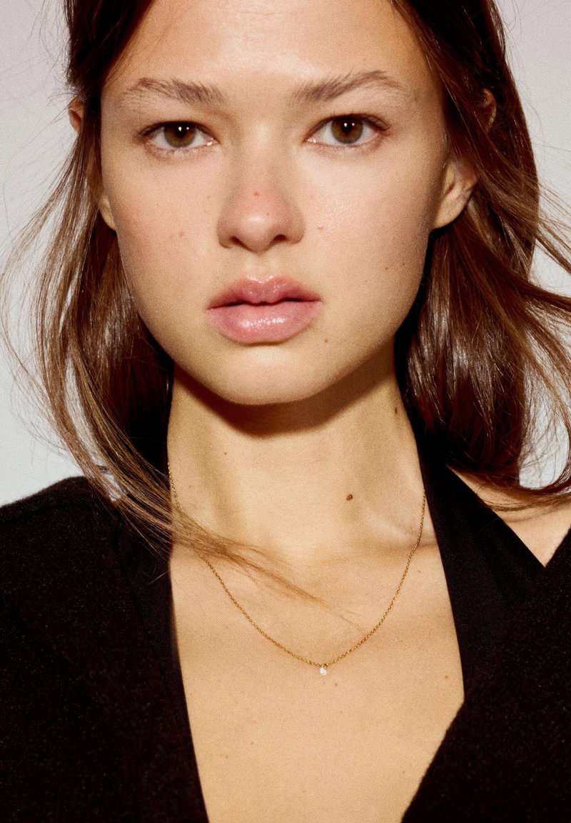 Jeune femme aux cheveux châtain clair, expression neutre, portant un fin collier en or et un haut noir sur un fond clair uni.