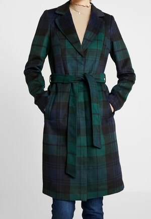 Femme portant un manteau ceinturé à carreaux verts et bleu marine jusqu'aux genoux, par-dessus un haut beige et un jean bleu, mains dans les poches du manteau.