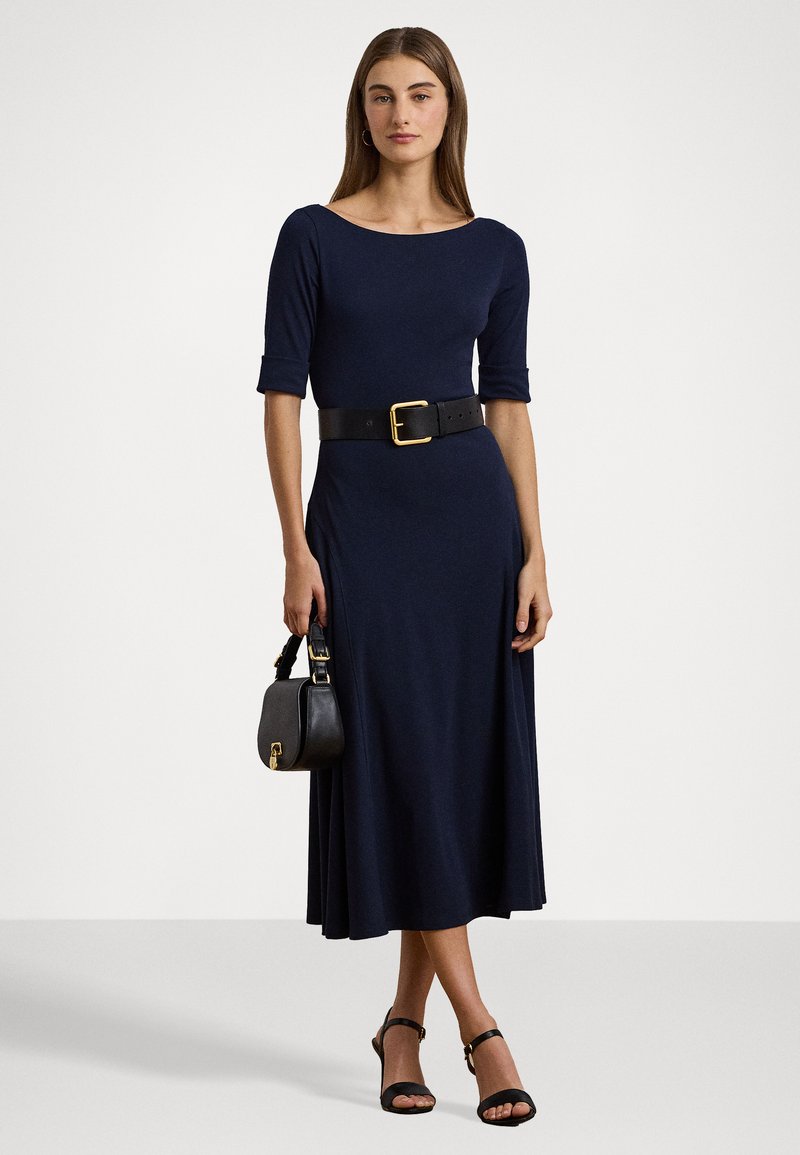 Robe bleu marine à manches trois-quarts avec un corsage ajusté, jupe fluide, ceinture en cuir noir avec boucle dorée, et petit sac à main noir.