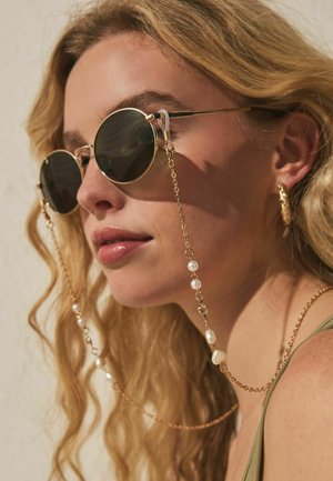 Femeie cu păr blond ondulat îi poartă ochelari de soare rotunzi și închiși, atașați unei lanț de aur cu accente de perle, cercei aurii în formă de cerc și un top verde.