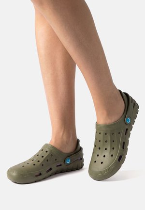 Pies con zuecos verdes oliva de tipo slip-on, con agujeros de ventilación y correas ajustables en el talón sobre un fondo blanco.