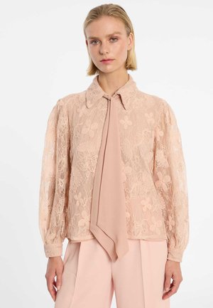 Vrouw die een lichtroze kanten blouse met bloemmotieven en een brede das draagt, gecombineerd met bijpassende lichtroze broek.