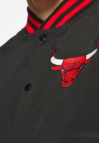 Schwarze Jacke mit einem rot-gestreiften, gerippten Kragen, ausgestattet mit einem roten gestickten Bullenlogo auf der linken Brust und schwarzen Druckknöpfen.