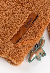 Braun flauschiger Fleece-Stoff mit einer strukturierten Oberfläche, versehen mit einem Lederetikett und einem stofflichen Etikett in Form eines Fuchses.