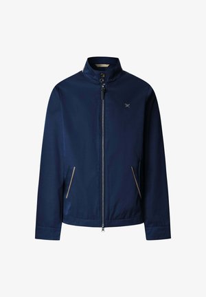 Veste zippée bleu marine avec col montant, deux poches avant avec bordure beige et petit logo blanc de clubs de golf croisés sur la poitrine.