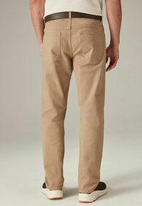 Beige katoenen broek met twee achterzakken, platte naden en een bruine riem. Valt nauw langs de benen en is gecombineerd met donkere sneakers.