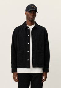 Μαύρο τζιν μπουφάν με διακριτικό λουλουδάτο σχέδιο, γιακά, κουμπιά μπροστά και πλαϊνές τσέπες. Φορεμένο πάνω από λευκό πουκάμισο και μαύρο παντελόνι.