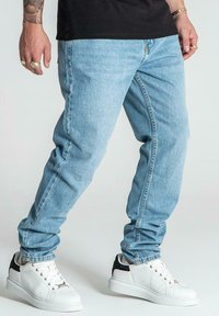 Vaqueros de mezclilla azul claro con un ajuste relajado, costuras visibles y un acabado desgastado. Combinados con zapatillas blancas con acentos negros.