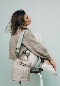 Beige leren emmer tas met trekkoord sluiting en lichtgroene verstelbare schouderband. Model draagt een gestructureerde bruine trui en witte gebreide broek.
