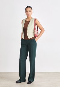 Gilet verde senza maniche con bottoni, top marrone, pantaloni verde scuro e una borsa rossa con motivo. Tessuti lisci, taglio sartoriale e tasche laterali.