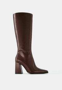 Bottes montantes en cuir marron avec un bout pointu et un talon épais. Le talon est orné d'un détail transparent pour plus d'élégance. Texture lisse.