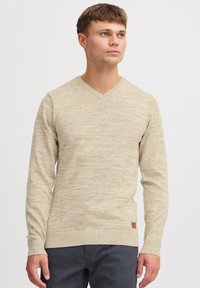Beige v-kaulusneule, joka on valmistettu teksturoidusta neulekangasta ja jossa on pitkät hihat. Siinä on ribbireuna ja pieni nahkalogo aivan alaosassa.