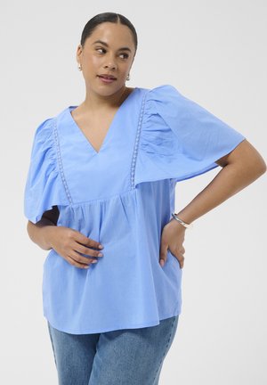 Donna con capelli scuri tirati indietro indossa una maglietta azzurro chiaro a scollo a V con maniche larghe arricciate e jeans blu, posando con la mano sul fianco.