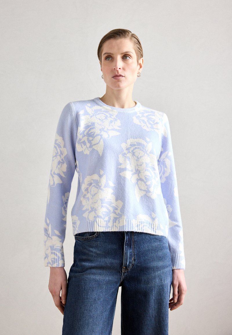 Lys blå genser med hvitt blomstermønster, ribbestrikket halskant og kant, kombinert med mørkeblå denimjeans. Myk tekstur, avslappet passform.
