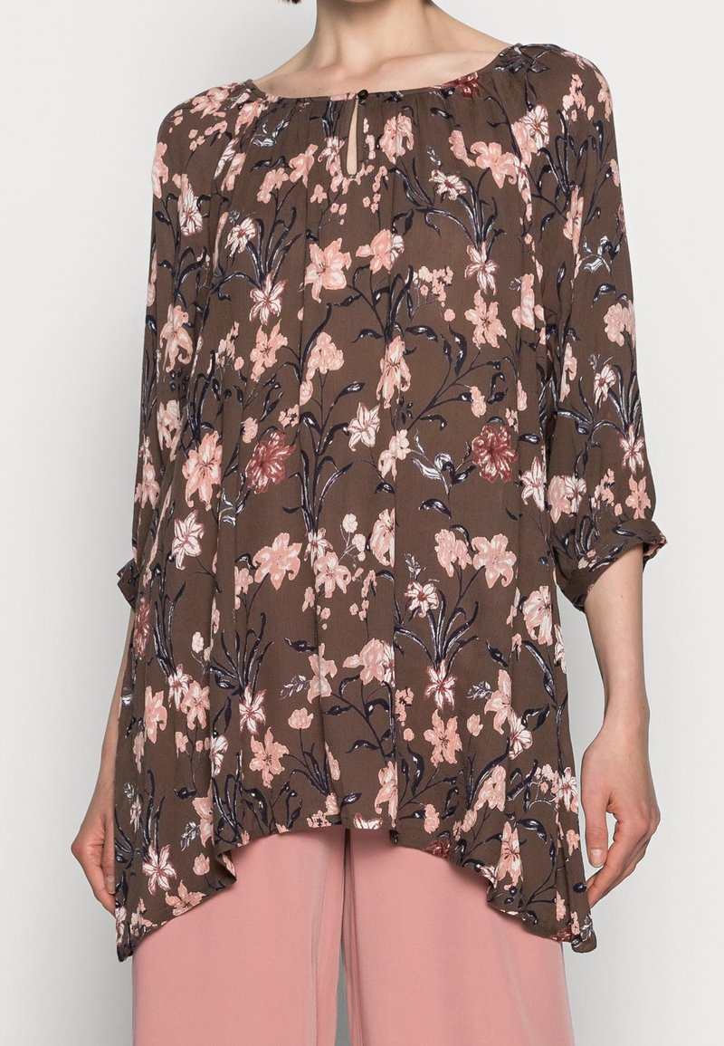 Blouse marron à motif floral avec des fleurs roses et violettes, manches longues, coupe ample et encolure en forme de goutte, portée avec un pantalon rose clair.