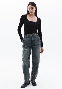 Schwarzes Langarmoberteil mit quadratischem Ausschnitt, kombiniert mit hochsitztenden, ausgewaschenen blauen Jeans. Das Modell trägt schwarze Slip-On-Schuhe.