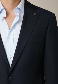 Granatowa marynarka o gładkiej fakturze, z jednym guzikiem i kieszenią na piersi. Pod spodem jasnoniebieska koszula z kołnierzykiem, z białymi guzikami.