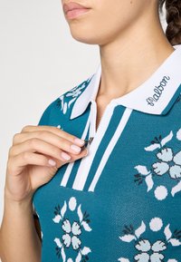 Teal strikke polo skjorte med blomstermønster, hvit krage og glidelås, med svarte detaljer og brodert merkevare på kragen.