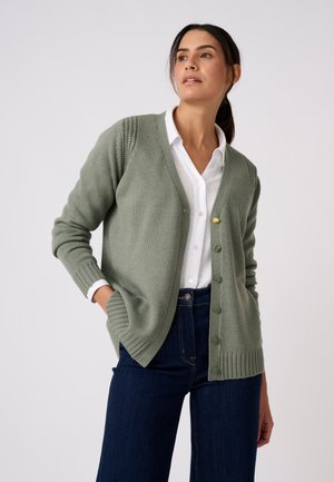 Vrouw draagt groene gebuttonde cardigan over wit hemd en donkerblauwe spijkerbroek, poseert met handen in de zakken tegen een effen achtergrond.