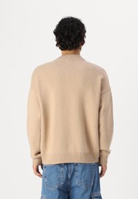 Beżowy sweter dzianinowy z ribbowym wykończeniem u dołu i przy mankietach, o luźnym kroju i długich rękawach. Tył prezentuje czysty, minimalistyczny design.