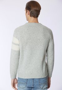 Maglione grigio girocollo in un tessuto morbido e testurizzato. Presenta strisce bianche sulle maniche. Indossato con jeans di denim blu.