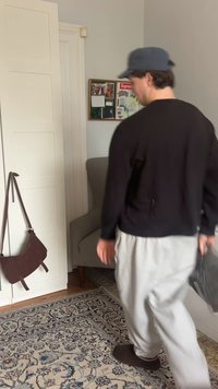 Férfi fekete pólóban és szürke nadrágban sétál egy szobában, amelyben mintás szőnyeg, szürke karosszék, barna táska és hirdetőtábla található a falon.