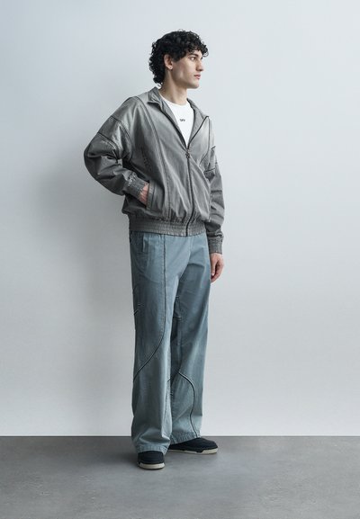 Νεαρός άνδρας με σγουρά μαλλιά που φοράει γκρι μπουφάν με φερμουάρ, λευκό πουκάμισο, φαρδιά μπλε παντελόνια με μαύρες ρίγες και μαύρα αθλητικά παπούτσια, στέκεται πλαγίως.