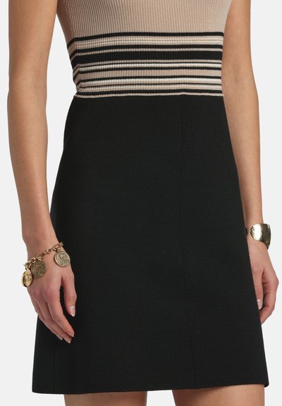 Fekete kötött ruha, bézs és fekete csíkos bordázott felsőrésszel. Alkalmazkodó derékkal és egyenes, A-vonalú szoknyával rendelkezik. Arany lánc karkötő a csuklón.