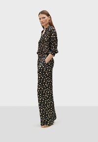 Black patterned jumpsuit with large beige spots, featuring long sleeves, a collar, and pockets. Smooth fabric with a relaxed fit.