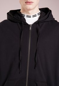 Felpa nera con zip in tessuto testurizzato e cappuccio con coulisse. Presenta una zip argentata e dettagli a coste all'orlo e al collo.