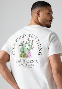Hvit bomull T-skjorte med et fargerikt kaktusgrafikk på baksiden, med teksten "Det er en vill vest ting" og "California Cacti Club."