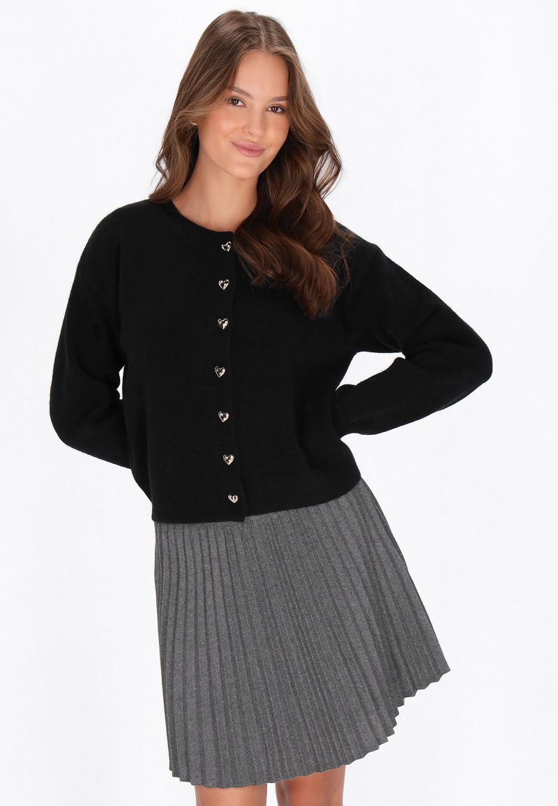 Svart stickad cardigan med hjärtformade knappar, kombinerad med en grå plisserad kjol. Mjuk textur, avslappnad passform och rund halsringning.