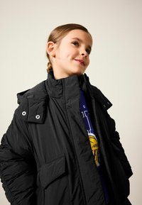 Schwarze Pufferjacke mit hohem Kragen, Druckknopfakzenten und zwei Fronttaschen; getragen über einem blauen Grafik-Sweatshirt.