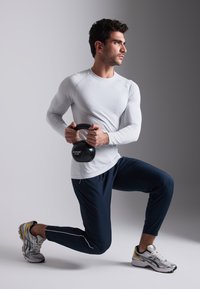 Man in a fitted white long-sleeve shirt and navy athletic pants, holding a 10 kg black kettlebell. Gray sneakers with yellow accents.