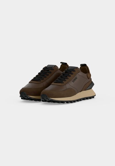 Paire de baskets basses marron avec des lacets noirs, des semelles beiges et des semelles extérieures noires texturées sur un fond uni clair.