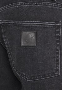 Zadnji žep črnih denim hlač s prišitim logotipom Carhartt na usnjenem označevalniku v usnjeni barvi, ki se ujema.